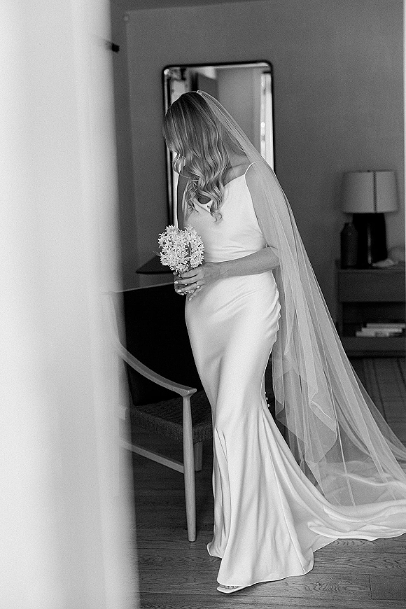 Bridal portrait in black and white of a bride holding bouquet, looking down in a hotel room by a mirror, long veil trailing behind
