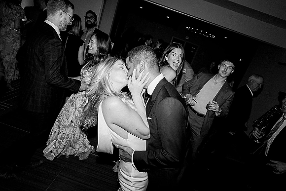 Wedding kiss moment as newlyweds kiss on the reception dance floor, bride in sleek dress and groom in tux, guests cheering under ceiling lights