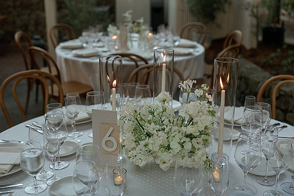 Reception tablescape with round wedding table setup, white floral centerpiece and taper candles in glass hurricanes on an outdoor patio with columns