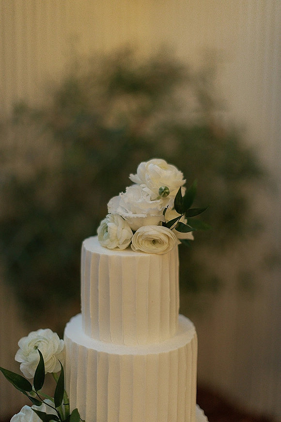Wedding cake with ribbed buttercream, two tiers topped with white sugar flowers and greenery against a soft neutral backdrop