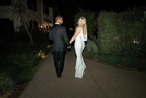 Couple portrait of bride and groom holding hands, walking down a garden path at night, bride in a low-back dress with veil looking back