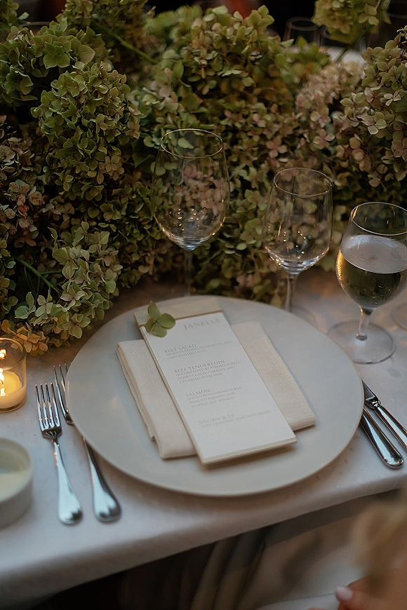 Reception tablescape with wedding place setting, menu card on a white plate, folded napkin, silver flatware, candles, and green hydrangea blooms