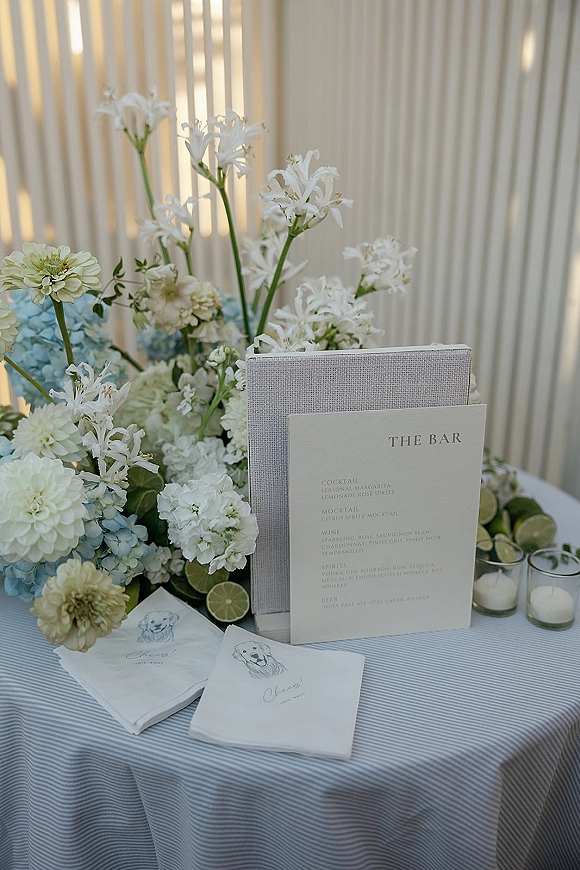 Wedding bar menu and signature cocktails sign on a striped tablecloth with white florals, blue hydrangea, limes, napkins, and votive candles