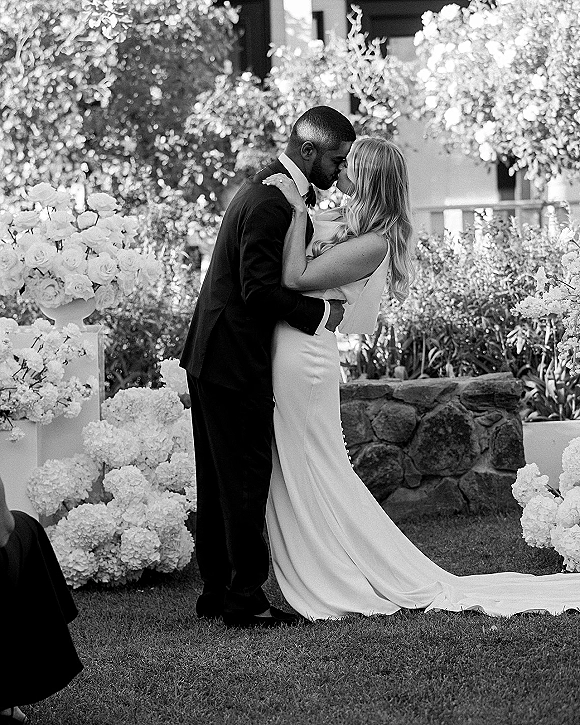 Wedding kiss portrait of bride and groom kissing, her strapless gown and train beside rose and hydrangea florals in a garden setting
