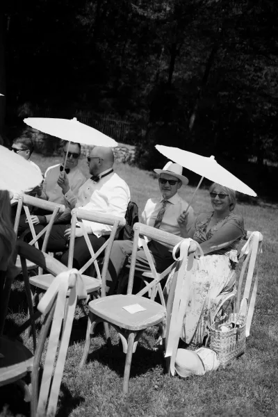 Ceremony guests shaded by wedding parasols for guests wait on a sunlit lawn with trees and a stone wall in the background.