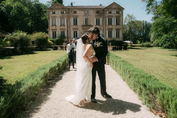 Wedding kiss portrait as the bride and groom kiss, her veil catching light before a historic mansion facade on a garden lawn.