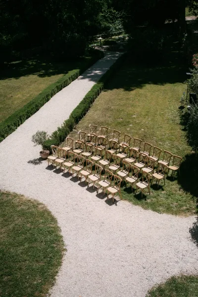 Ceremony seating for an outdoor ceremony setup with crossback chairs lining a gravel path beside a hedged walkway and lawn under garden trees.