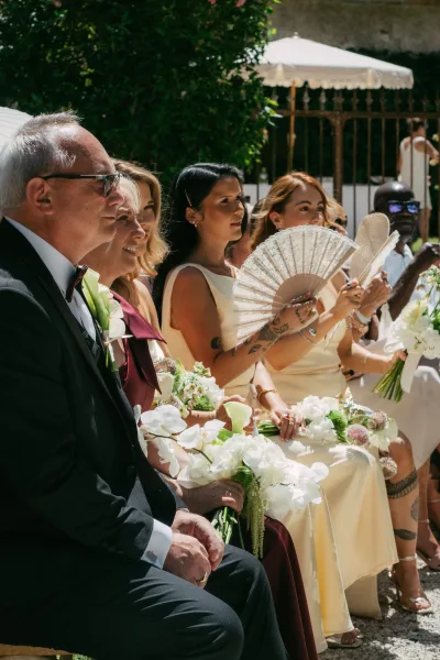 Ceremony guests at an outdoor wedding ceremony cool themselves with hand fans, seated by garden greenery and an iron fence on gravel ground.
