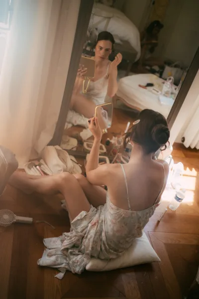 Bride getting ready pauses for a makeup touch up, studying her reflection in a handheld mirror, a calm, candid moment before the ceremony.