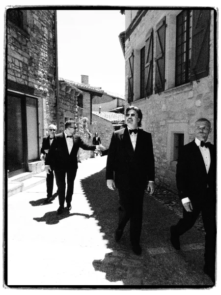 Groom and groomsmen stroll as black tie groomsmen down a sunlit cobblestone alley, wearing sunglasses amid stone buildings and shutters.