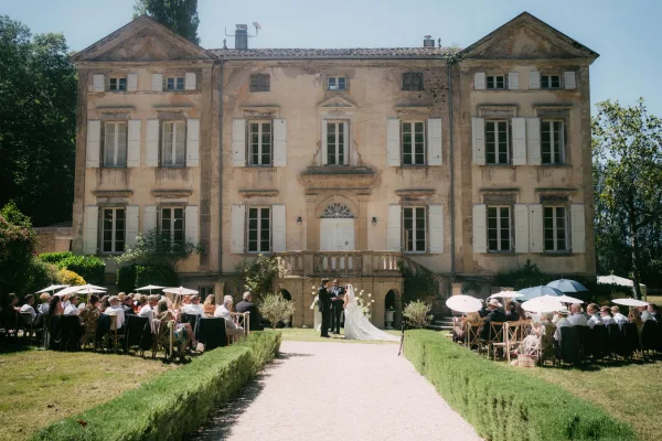 Outdoor ceremony setup with garden ceremony seating shaded by white parasols, arranged on a lawn before a historic mansion facade under blue sky.
