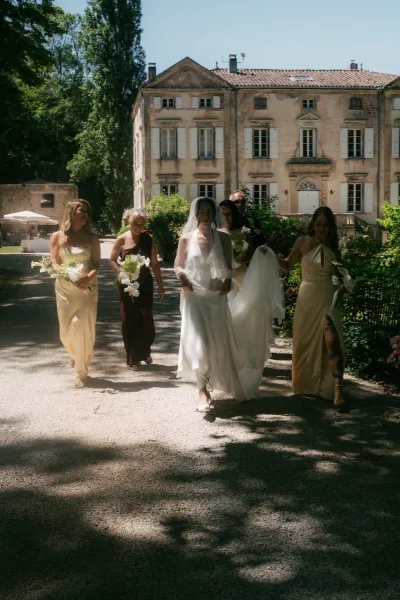 Bride with bridesmaids walking together, her sheer veil trailing on a gravel path by a historic manor and sunlit garden trees.