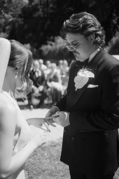 Ceremony ring exchange as the couple are exchanging wedding rings; groom slides a band onto her hand, bride’s veil catching light in a garden.