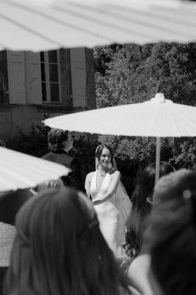 Ceremony moment at an outdoor wedding ceremony as the bride and groom face each other, guests shaded by paper parasols beneath garden trees.