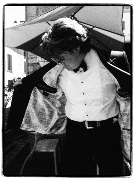 Groom portrait during groom getting ready, adjusting his tuxedo jacket on a patio by a stone building under a patio umbrella.