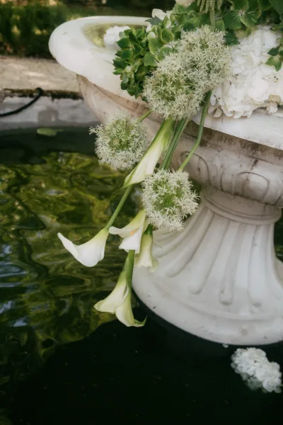 Fountain floral installation of wedding fountain flowers with calla lilies, framed by water and garden shrubs beside a gravel path.