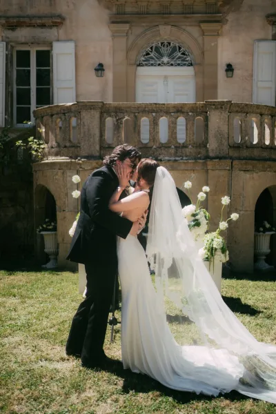 Wedding kiss portrait capturing the ceremony first kiss as the bride holds the groom, who wears sunglasses, before a stone facade and balustrade.