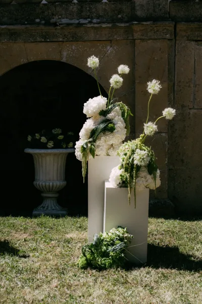 Ceremony floral arrangement with pedestal wedding flowers featuring white hydrangeas, set against a stone archway and grass lawn backdrop.