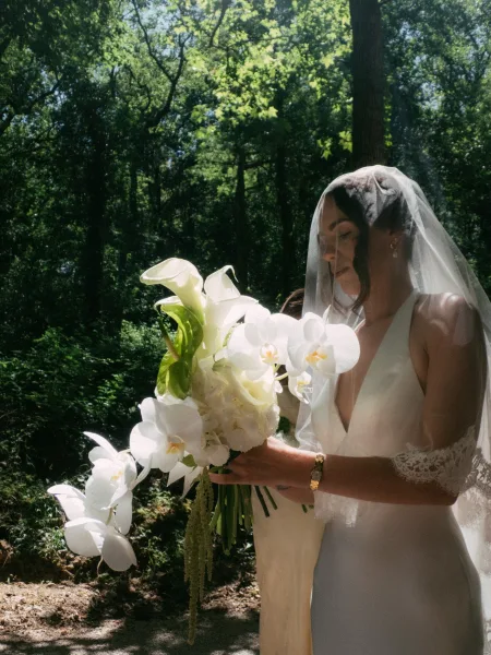 Bridal portrait of a bride cradling a white orchid bouquet on a dirt path amid forest trees dappled with sunlight and deep green leaves.