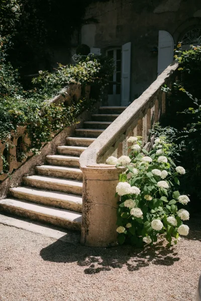 Stone staircase serving as a garden staircase backdrop, lined with white hydrangeas, against a stucco wall with open shutters and doorway.