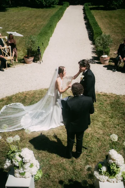 Ceremony moment as wedding vows are exchanged on a garden lawn beside a gravel aisle, framed by hedges and trees, the bride’s long veil trailing.