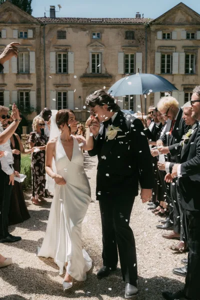 Confetti exit during the ceremony recessional as the bride and groom walk a gravel aisle, her simple veil trailing, before a historic mansion facade.