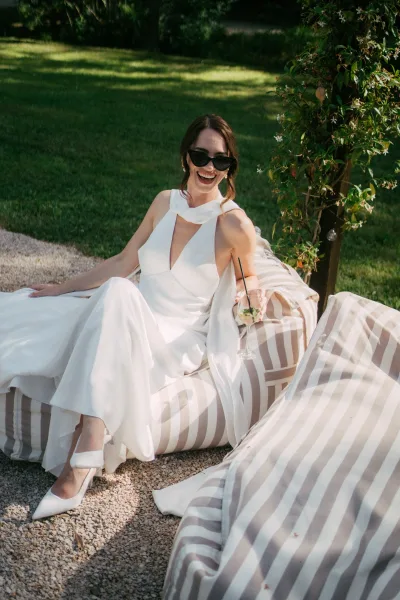 Bridal portrait of a bride with sunglasses relaxing in a garden beneath a wooden arbor draped with climbing vines.