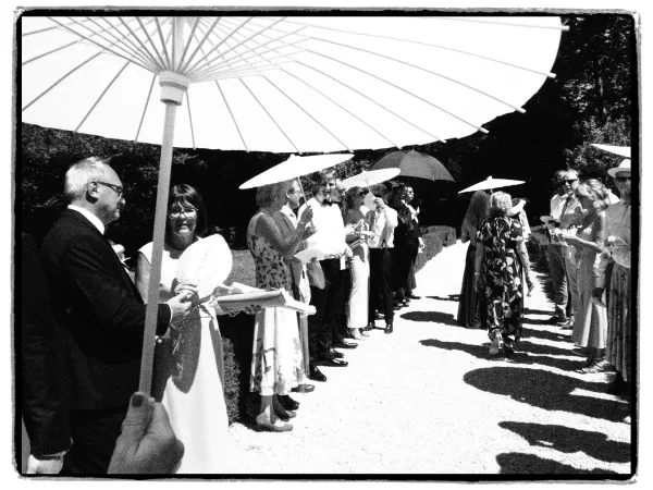 Ceremony moment as wedding guests with parasols line a gravel path amid garden trees and hedges, forming an aisle.