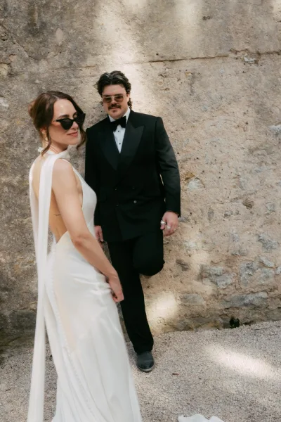 Couple portrait featuring a bride in sunglasses as she looks over her shoulder, with the groom beside her against a weathered stone wall.