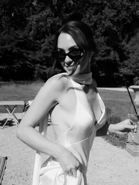 Bridal portrait of a bride in sunglasses, posing on a garden lawn framed by trees and a gravel path, with outdoor tables blurred in the background.