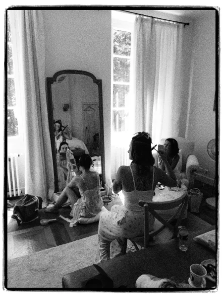 Bridal party getting ready as bridesmaids chat and style hair with a curling iron, sharing a candid, cozy pre-ceremony moment.