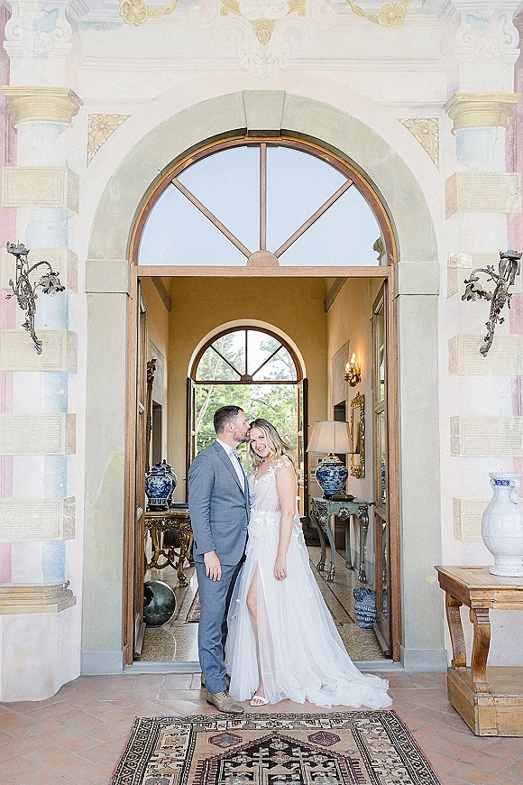Couple portrait of groom kissing bride’s forehead in a villa entryway, her veiled gown and heels framed by an arched doorway