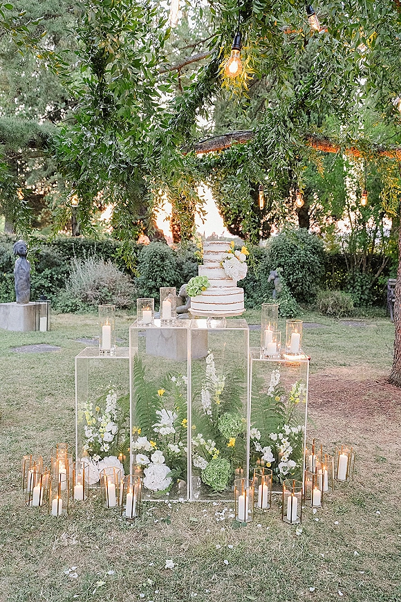 Wedding cake display with tiered cake on clear acrylic stands, surrounded by pillar candles and florals on a garden lawn under string lights