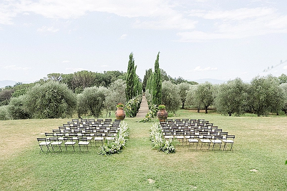 Ceremony setup for an outdoor wedding ceremony with wooden folding chairs, aisle florals and terracotta urns on a lawn among olive trees
