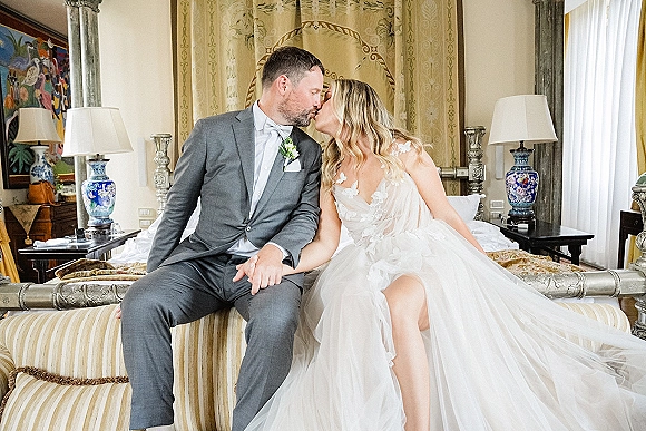 Wedding kiss portrait of bride and groom kiss on a bed, her lace bodice and tulle skirt beside him in a bow tie in a cozy bedroom suite