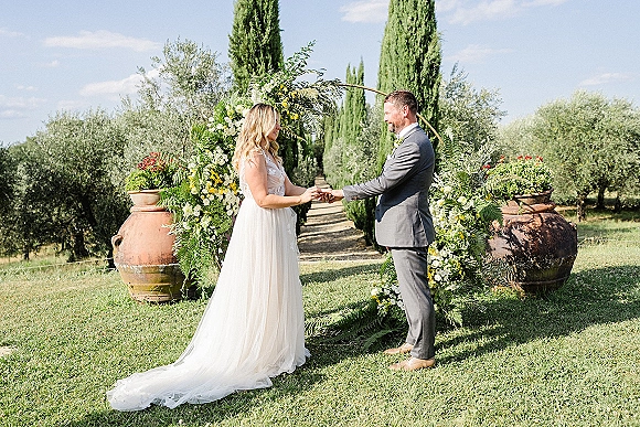 Wedding vows as bride and groom exchange rings under a floral arch with greenery, on a sunny lawn lined with cypress trees