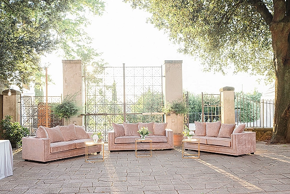 Wedding lounge seating with blush velvet sofas and gold side tables, styled with floral arrangements on a stone patio by a wrought iron gate
