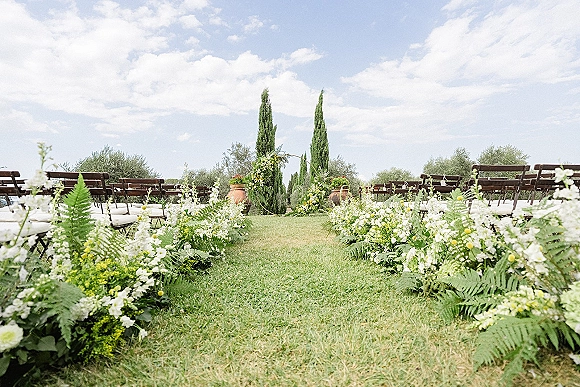 Ceremony aisle decor with ground floral arrangements of white and yellow blooms, ferns, and greenery leading to a circular arch on a lawn