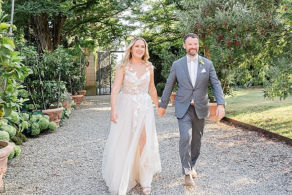 Newlywed couple portrait of bride and groom walking hand in hand on a garden path, lace dress visible, greenery and iron gate behind