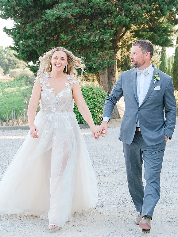 Couple portrait of bride and groom walking hand in hand, bride in lace dress and tulle skirt, on a tree-lined gravel path