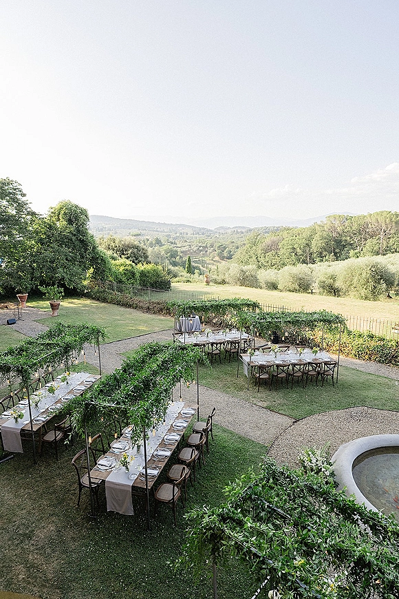 Outdoor reception setup with long white linen banquet tables and cross back chairs beneath greenery canopy and string lights on an estate lawn