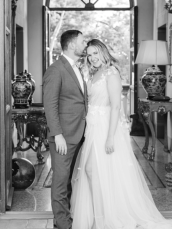 Couple portrait of bride in lace tulle gown smiling as groom kisses her forehead in a doorway with string lights and window trees behind