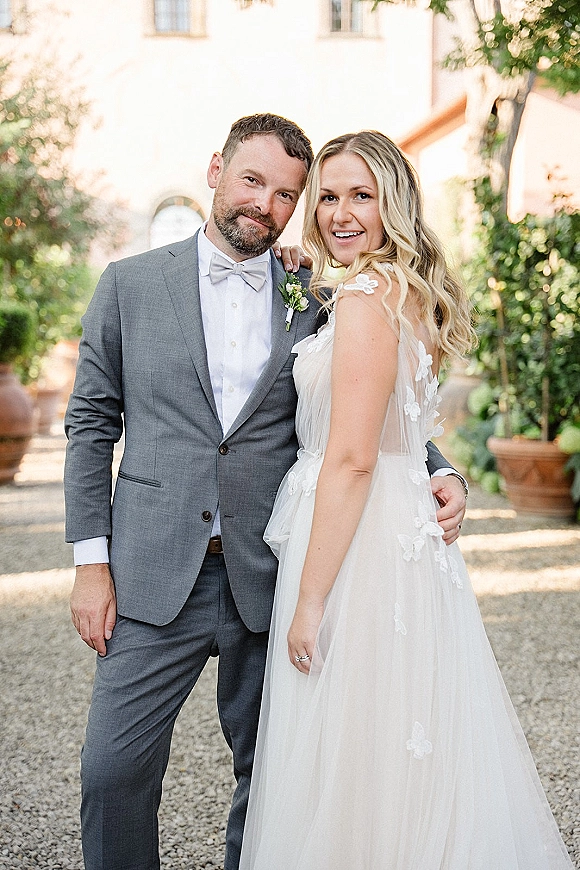 Couple portrait of bride and groom portrait, bride leaning on groom in a courtyard with greenery, floral appliqué gown and boutonniere visible