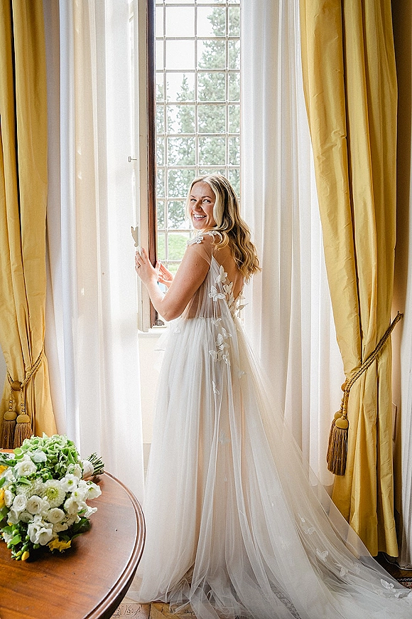 Bridal portrait of a smiling bride by window in a lace appliqué gown with long train, holding a bouquet in soft daylight by curtains