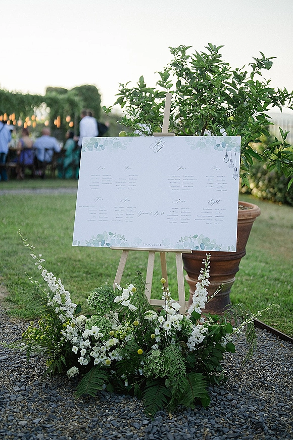 Wedding seating chart on a wooden easel with green-and-white floral ground arrangement and potted tree by an outdoor lawn reception under string lights