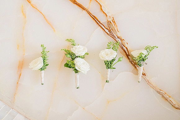 Wedding boutonnieres with white flowers and greenery tied in ribbon wraps, arranged as a flat lay on a marble surface