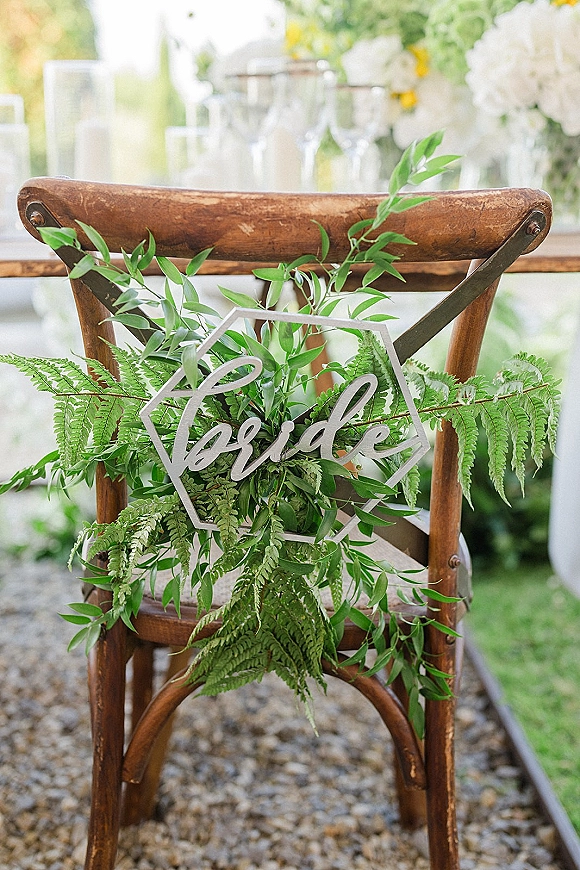 Wedding chair decor with a bride chair sign on clear acrylic, draped in fern greenery garland over a wood chair on an outdoor lawn aisle