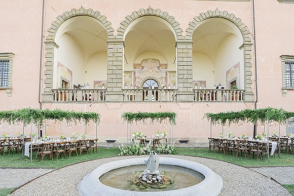 Outdoor reception setup with long banquet tables, white linens and greenery runners, under string lights in a courtyard with fountain