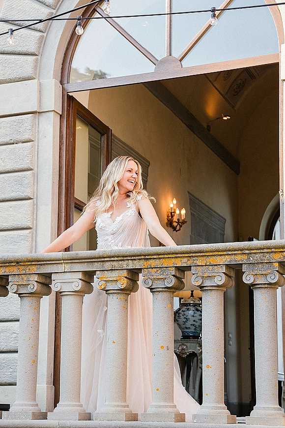 Bridal portrait of a bride on balcony, smiling in a sheer tulle gown with lace bodice, framed by columns and string lights near an arched doorway