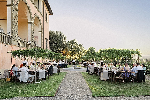 Outdoor wedding reception with long banquet tables and white linens under string lights and a greenery canopy beside an arched villa at sunset